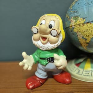 Può includere: Statuetta vintage di un personaggio dei cartoni animati, con una camicia verde, pantaloni grigi e scarpe rosse. La figura ha barba bianca, occhiali e un cappello giallo. Un mappamondo è sullo sfondo.