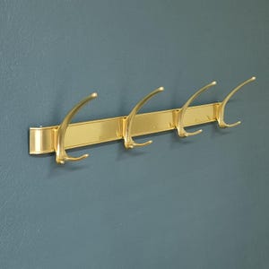 Op de afbeelding: Een goudkleurige wandgarderobe met vier gebogen haken. De haken zijn bevestigd aan een rechthoekige balk, allemaal in een bijpassende gouden afwerking. De garderobe is gemonteerd op een blauwgroene muur.