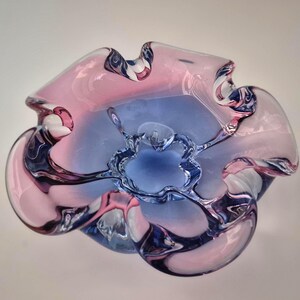 Könnte beinhalten: Eine dekorative Glasschale mit einer einzigartigen, organischen Form. Die Schale hat eine Mischung aus rosa, blau und klarem Glas, wodurch ein optisch ansprechender Farbverlauf entsteht. Das Design umfasst fließende Kurven und eine zentrale, blütenartige Form.