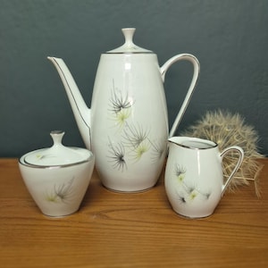 Peut inclure: Service à thé en céramique blanche comprenant une théière, un sucrier et un pot à crème. Chaque pièce est ornée d'une bordure argentée et d'un motif floral noir et vert-jaune. L'ensemble est présenté sur une surface en bois.