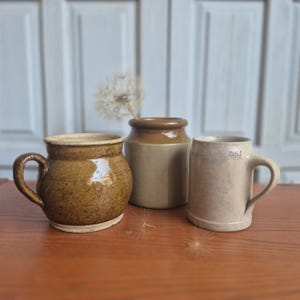 Peut inclure: Un ensemble de trois récipients en céramique comprenant une tasse ronde marron mouchetée avec une anse, un pot cylindrique beige avec un bord marron foncé et une tasse gris clair avec une anse portant l'inscription 0.5L sur le côté.