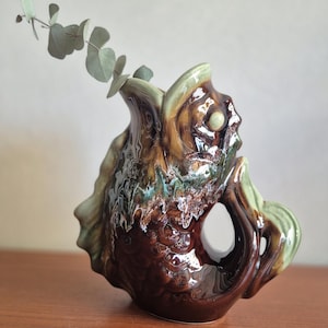 May include: A glazed ceramic pitcher shaped like a fish with a dark brown body, light green fins, and a light green rim. The pitcher features a textured scale pattern and a handle formed by the tail. A small sprig of eucalyptus is inside the opening.