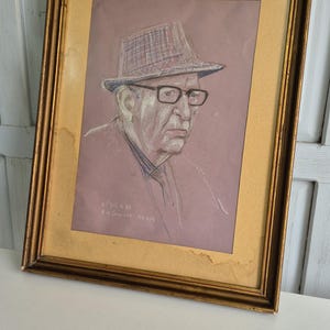 May include: A framed portrait of an elderly man wearing spectacles and a checked hat. The artwork is a sketch with a gold-coloured frame. The background is a light purple colour. The artwork is signed in the bottom left corner.