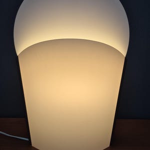 May include: A modern, illuminated table lamp with a unique design. The lamp features a white, rounded top and a cream-colored, trapezoidal base. The lamp is made of a translucent material, and the light source is visible from the bottom.