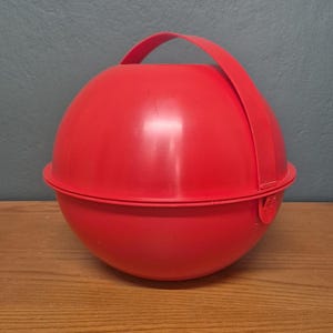 Peut inclure: Un récipient sphérique rouge vif avec une poignée assortie. Le récipient est fait d'un matériau lisse et solide et présente une couture au milieu. La poignée est fixée au sommet de la sphère. Le récipient est posé sur une surface en bois.