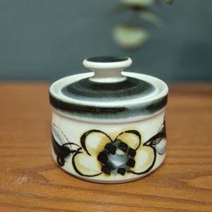 Peut inclure: Un petit sucrier en céramique avec un couvercle. Le bol est blanc avec un motif floral noir, jaune et gris. Le couvercle est blanc avec une bande noire et grise. Le sucrier est sur une surface en bois.