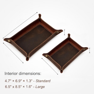 May include: Two brown leather catchall trays with snap closures. The larger tray measures 6.5" x 8.5" x 1.6", and the smaller tray is 4.7" x 6.9" x 1.3". The trays have a rectangular shape with raised edges and are designed to hold small items.