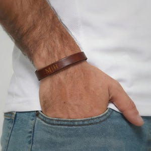 Op de afbeelding: Een bruine leren armband met de initialen "M|H" gegraveerd op de voorkant. De armband wordt om een pols gedragen en de persoon draagt een wit shirt en een blauwe jeans.