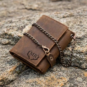 May include: A brown leather tri-fold wallet featuring a metal chain attached to a side loop. The front of the wallet is embossed with the letters MR centered between two stylized antlers. The leather has a distressed finish with visible stitching.