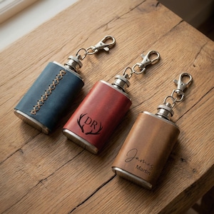 Op de afbeelding: Drie met leer beklede heupflessen met sleutelhangers. Eén is blauw met stiksels, een andere is rood met "DR" en geweien, en de derde is bruin met "James Martin". Allemaal hebben ze zilveren fittingen.