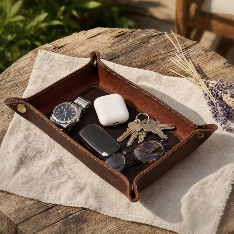 May include: A brown leather catchall tray with a watch, keys, wireless earbuds, car key fob, and eyeglasses. The tray is on a linen cloth on a wooden table. The tray has stitched edges and brass rivets.