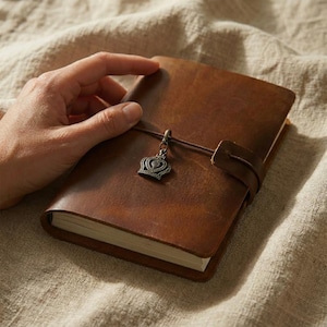 May include: A brown leather journal with a smooth, matte finish and a wrap-around leather cord closure. A small, antique-style bronze metal crown charm hangs from the cord on the front cover. The journal contains white paper pages.