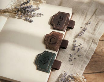 May include: Three leather pen holders with elastic bands attached to the edge of an open notebook. The top holder is dark brown with the letters M and K separated by a line. The middle is brown with a C inside a wreath. The bottom is dark green with a butterfly.