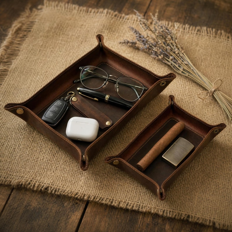 May include: Two brown leather catchall trays with items inside. The larger tray holds eyeglasses, a pen, car keys, and a white case. The smaller tray contains a cigar and a lighter. A bundle of dried lavender is in the background.