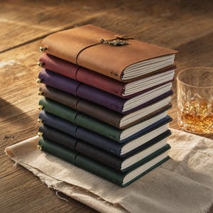 May include: A stack of seven rectangular journals with matte covers in tan, maroon, purple, brown, green, navy, and black. Each journal features white pages, a vertical elastic closure, and a small metallic charm attached to the front cover.