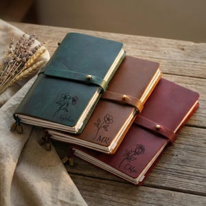 May include: Three rectangular leather journals in dark green, brown, and burgundy. Each features a wrap-around strap with a brass stud closure, a laser-engraved floral illustration, and a personalized name: Sophia, MR, and Chloe, respectively.