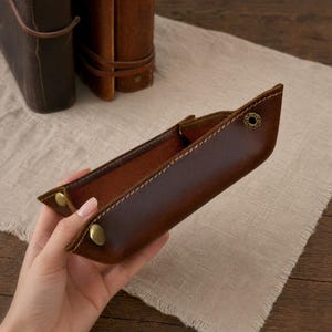 May include: A brown leather tray with brass-colored snap closures. The tray is rectangular with a stitched border and a lighter brown interior. Two leather journals are in the background.