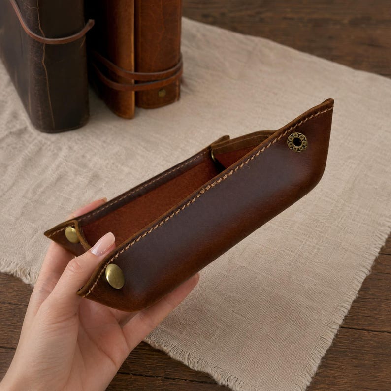 May include: A brown leather tray with brass-colored snap closures. The tray is rectangular with a stitched border and a lighter brown interior. Two leather journals are in the background.