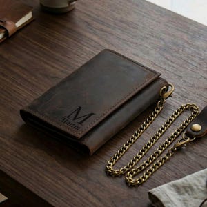 May include: A dark brown leather wallet featuring a distressed finish and a metal chain attachment. The front of the wallet is engraved with a large letter M and the name Martin below it. The wallet has a rectangular shape and visible stitching.