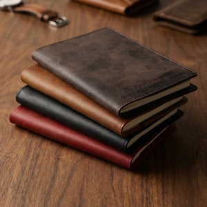 Op de afbeelding: Een stapel van vier leren dagboeken in donkerbruin, tan, zwart en rood. Het bovenste donkerbruine dagboek bevat de tekst in reliëf: This journal holds secrets, dreams, and everything in between. Elk dagboek heeft een matte afwerking.