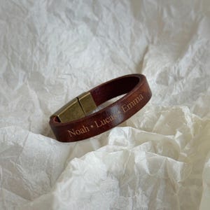 May include: A dark brown leather bracelet featuring a metallic bronze-toned magnetic clasp. The surface of the leather band is engraved with the names Noah, Lucas, and Emma, separated by small dots, creating a personalized accessory design.