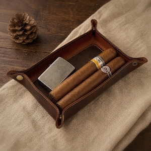 May include: A brown leather tray with a lighter and two cigars. The cigars have yellow and white bands. The tray is rectangular with rounded corners and is on a tan surface.