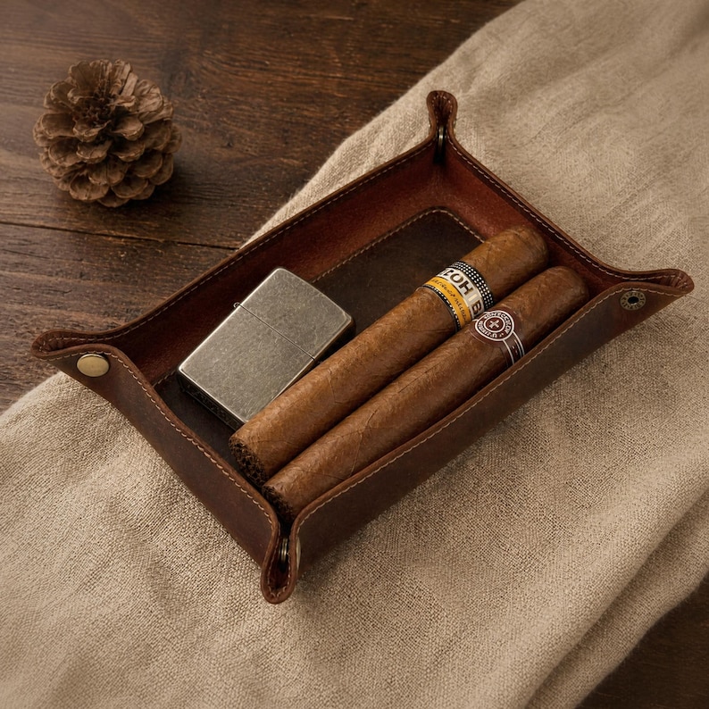May include: A brown leather tray with a lighter and two cigars. The cigars have yellow and white bands. The tray is rectangular with rounded corners and is on a tan surface.