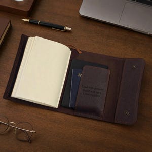 May include: A dark brown leather journal cover with a snap closure, containing a notebook and a passport holder. The interior pocket features the embossed text: Lead with purpose. Build with care. Grow together. A black fountain pen sits nearby.