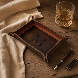 May include: A brown leather valet tray with the initials "TAT" and the name "MARTIN" engraved inside. The tray is on a wooden surface, with a glass of amber liquid and a pen nearby.