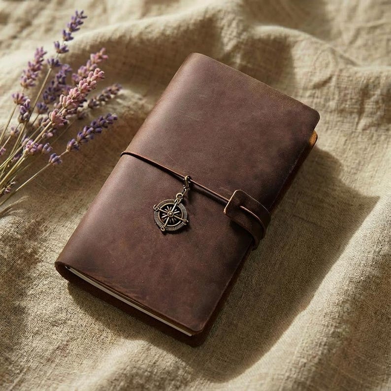 May include: A dark brown leather journal with a wrap-around strap closure. A metallic bronze-toned compass charm is attached to the strap. The journal is rectangular with a smooth, matte finish and visible stitching along the edges of the cover.