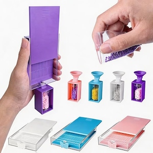 Op de afbeelding: Een set opbergtools voor diamond painting met rechthoekige trays met gegroefde oppervlakken in wit, blauw en roze, en kleine doorzichtige glazen flesjes met kleurrijke trechterdoppen gevuld met paarse, gele en roze kraaltjes.