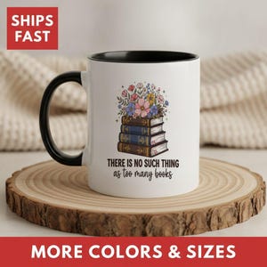 May include: A white ceramic mug with a black handle and interior rim. The front features a graphic of a stack of three books topped with colorful flowers and the text, "THERE IS NO SUCH THING as too many books" in black lettering.
