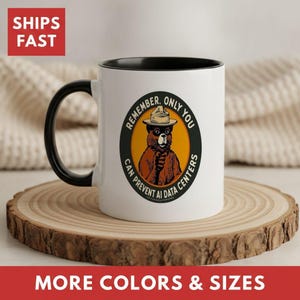 May include: A white ceramic mug with a black handle and interior rim. The front features a circular graphic of a brown bear wearing a ranger hat, with the text "REMEMBER, ONLY YOU CAN PREVENT AI DATA CENTERS" printed in black around the orange circle.