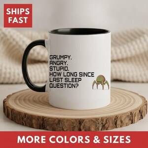 May include: A white ceramic mug with a black handle and interior. The mug features black text that reads: GRUMPY. ANGRY. STUPID. HOW LONG SINCE LAST SLEEP QUESTION? Next to the text is a small, green and brown illustration of a multi-legged creature.