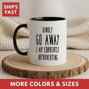 May include: A white ceramic mug with a black interior and a black handle. The mug features black text that reads: KINDLY GO AWAY I AM CURRENTLY INTROVERTING. The mug has a cylindrical shape with a smooth finish and a sturdy handle on the side.
