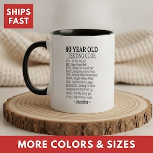 May include: A white ceramic mug with a black handle and interior, featuring black text that reads 80 YEAR OLD TEXTING CODE followed by a list of humorous acronyms and the name Jennifer printed in black on the front surface of the mug.