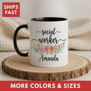 May include: A white ceramic mug with a black handle and interior rim. The front features the black text "social worker" above a colorful floral illustration of pink, orange, and green leaves, with the name "Amanda" printed in black below.