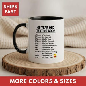May include: A white ceramic mug with a black handle and interior, featuring black printed text titled 65 YEAR OLD TEXTING CODE. The list includes humorous abbreviations like ATD for At The Doctor, BFF for Best Friend Fell, and ROFLACGU for Rolling On Floor Laughing And Can't Get up, accompanied by a laughing emoji.
