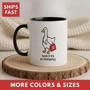 May include: A white ceramic mug with a black handle and interior rim. The mug features a cartoon illustration of a white goose wearing a stethoscope and carrying a red medical kit. Below the goose, the text reads: honk if it's an emergency.