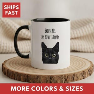 May include: A white ceramic mug with a black handle and interior rim. The front features a graphic illustration of a black cat with large yellow eyes, positioned below the black text that reads, Excuse Me, My Bowl Is Empty.