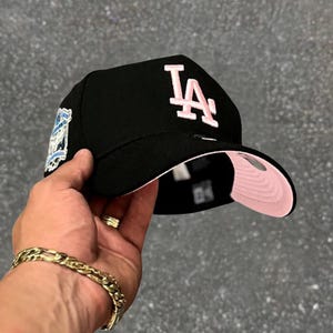 Può includere: Cappellino da baseball nero con le lettere "LA" in rosa sul davanti. Il cappellino ha una visiera inferiore rosa e una toppa sul lato. Il cappellino è tenuto in mano.