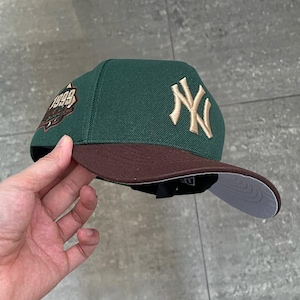 Può includere: Un cappellino da baseball verde con visiera marrone e logo "NY" dorato. Il cappellino presenta una toppa con l'anno "1999" sul lato. L'interno della visiera è bianco.