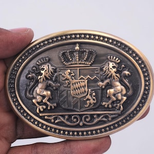 May include: An oval-shaped belt buckle with an antique brass finish featuring a central coat of arms. The design includes two rampant lions flanking a shield topped with a crown, all surrounded by a decorative beaded border and scrollwork.