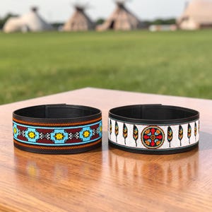 May include: Two black leather cuffs featuring intricate beadwork. The left cuff displays a geometric pattern in turquoise, yellow, and maroon. The right cuff features a central circular motif surrounded by vertical feather designs in white, red, and blue.