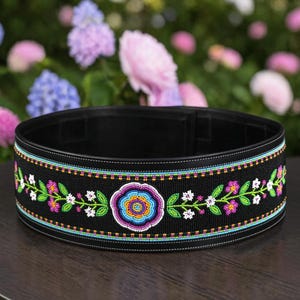 May include: A black belt with a beaded floral design. The belt features a central flower with a blue, purple, and yellow pattern, surrounded by green leaves and pink and white flowers. The belt is approximately 3 inches wide.