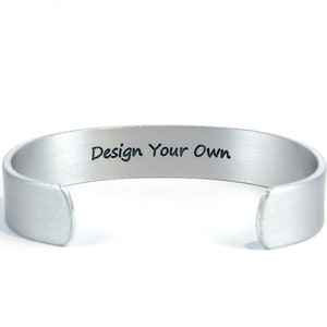 May include: Silver metal bracelet with a blank space for engraving. The bracelet has the words "Design Your Own" engraved on it.
