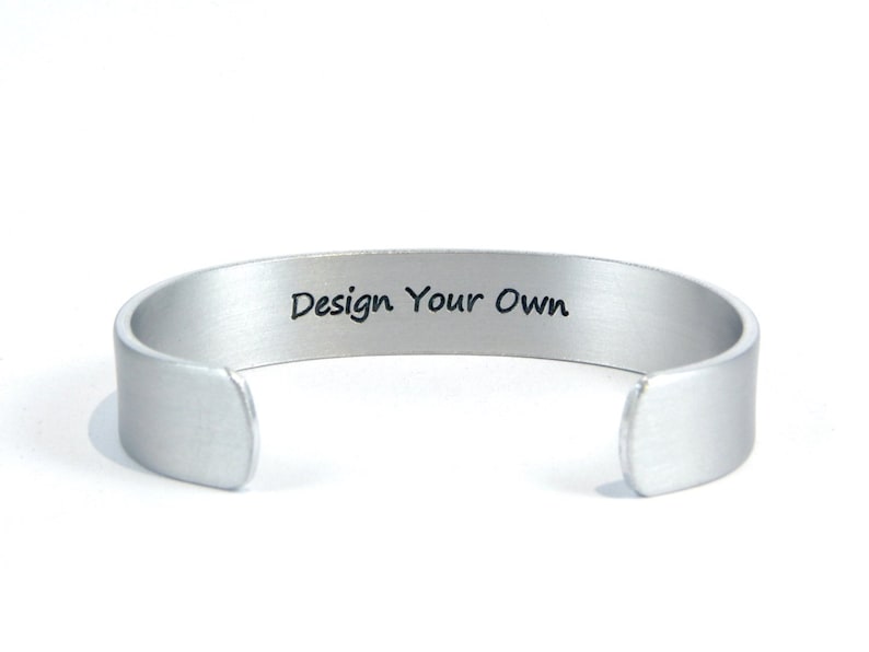 May include: Silver metal bracelet with a blank space for engraving. The bracelet has the words "Design Your Own" engraved on it.