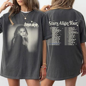 Puede incluir: Camiseta gris oscuro con una imagen en blanco y negro de Freya Skye. La parte delantera tiene el texto "freya skye". La parte trasera muestra "Stars Align Tour" y una lista de fechas y lugares de la gira, incluyendo ciudades europeas.