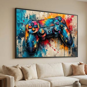 May include: A rectangular wall art print featuring a vibrant, abstract watercolor-style painting of a video game controller. The controller is rendered in bold splashes of blue, orange, red, and yellow paint against a textured, neutral background.