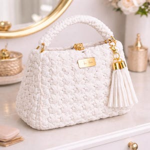May include: A white, crocheted handbag with a structured shape and a rounded handle. The bag features gold-toned hardware, including a clasp, a small tag, and a tassel. The bag is approximately 9 inches wide.